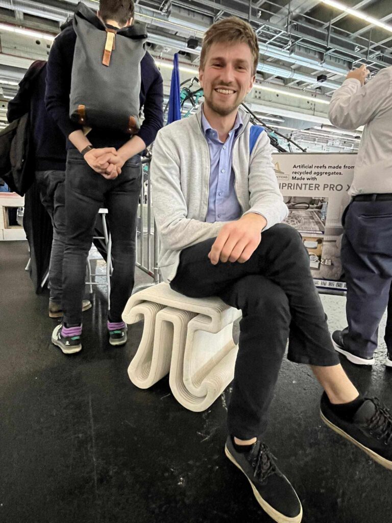 Martynas Karvelis lankosi formnext parodoje, dalinasi įspūdžiais apie betono 3D spausdinimą, sėdi ant betoninės 3D spausdinto kėdės